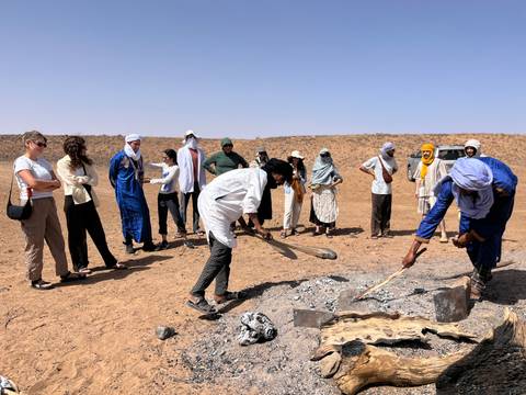 Tourists and Berber hosts gather around a sand oven baking flatbread in the open desert.