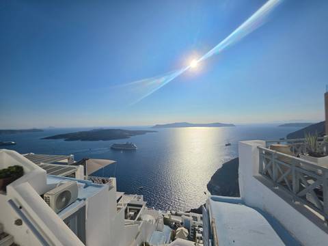 High noon sun glints over caldera waters with cruise ships anchored beside white cliff villages.