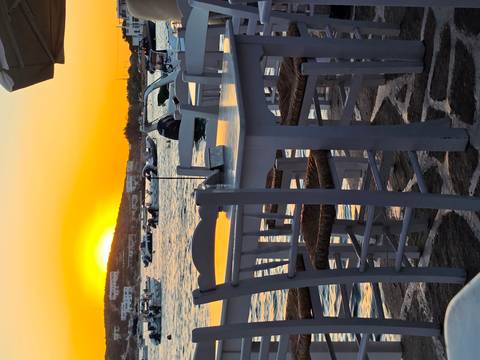 Empty waterfront taverna tables silhouetted against vibrant setting sun over harbour.