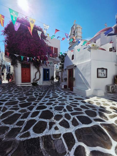 Charming Mykonos alley with stone mosaic pavement, bright doors and bougainvillea under bunting flags.