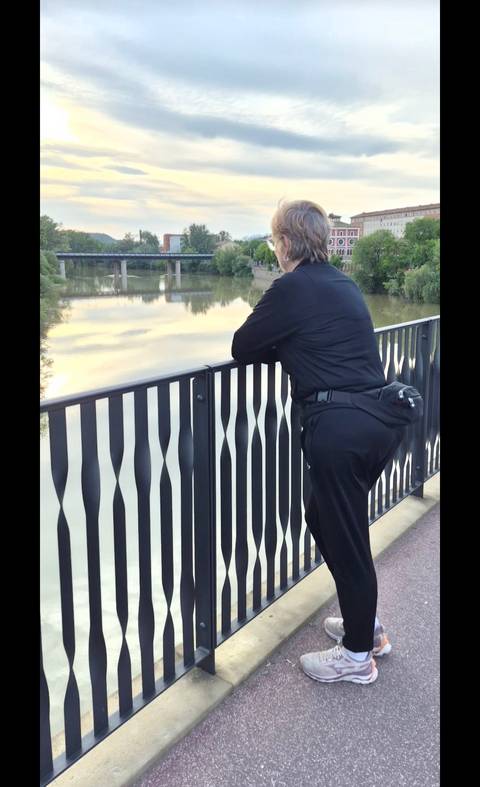 Person in black leisurewear leans on a patterned metal railing overlooking a calm river at dusk.