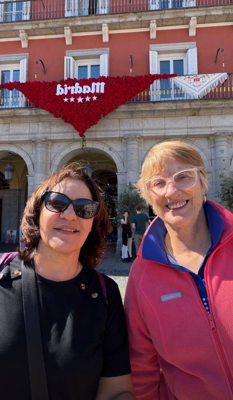 Two women smile for a selfie in front of a grand stone arcade in a sunny plaza.