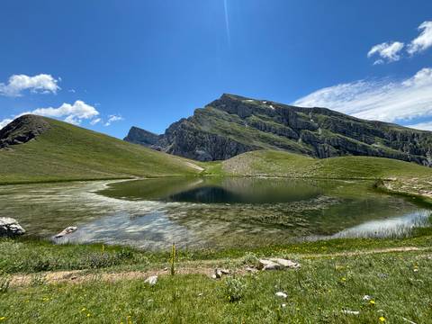 A crystal-clear alpine lake mirrors jagged green mountains under a bright blue sky.