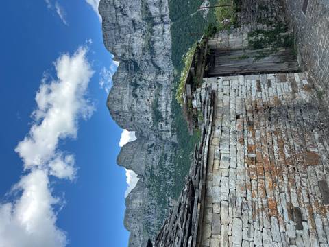 Stone wall foreground leads to towering limestone cliffs under a deep blue sky with small clouds.