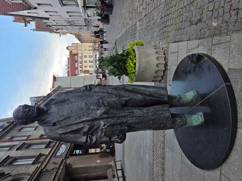 Bronze statue of a man in a long coat stands on a pedestrian street lined with historic buildings.