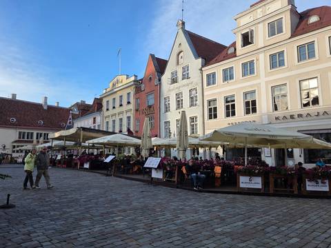 Outdoor cafés with umbrellas fill a medieval town square lined with pastel gabled buildings.