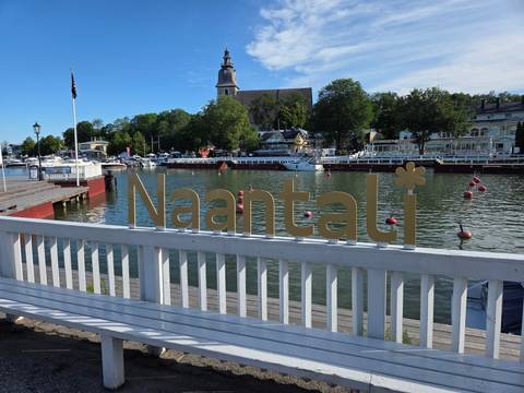 Golden Naantali sign mounted on a white railing overlooks a calm harbor with boats and trees.