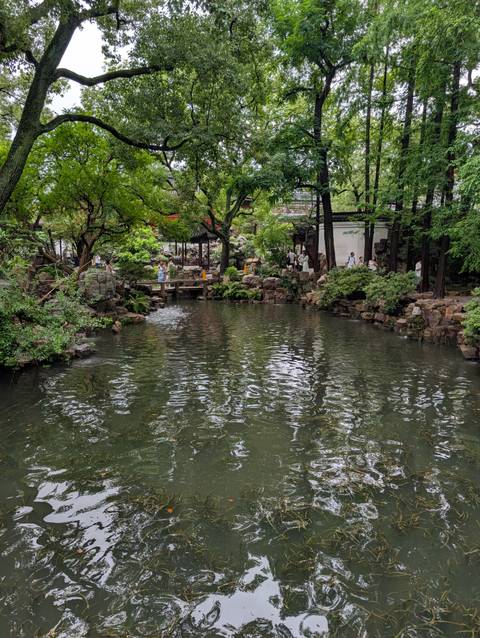 Lush classical Chinese garden with visitors walking bridges around a tranquil reflective pond.