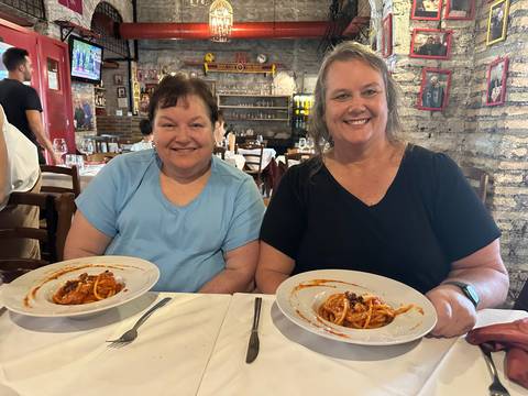 Two women smiling in a cozy brick-walled restaurant holding plates of pasta.