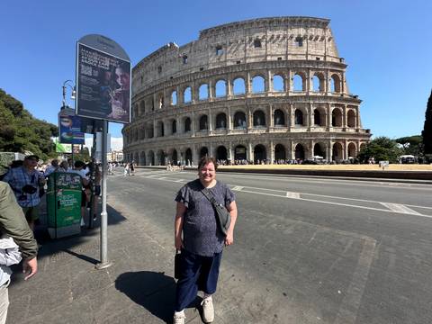 Woman standing on sidewalk with the Colosseum towering across the street under bright sun.
