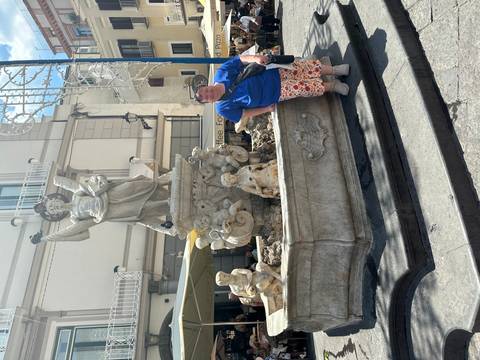 Woman leans on a stone fountain adorned with angel statue in a sunlit town square.