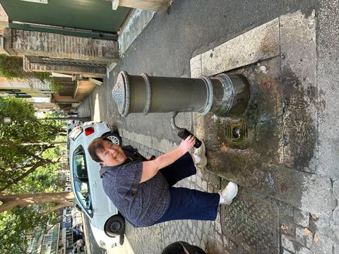 Visitor refilling a bottle at a traditional street fountain along a leafy urban lane with parked cars.