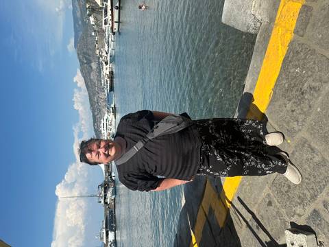 Visitor posing beside clear harbour water with yachts and a mountainous coastline behind.