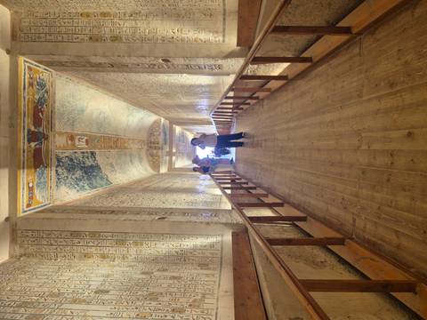 Decorated corridor with ancient hieroglyphs and colorful ceiling art inside a pharaonic tomb as visitors take photos.