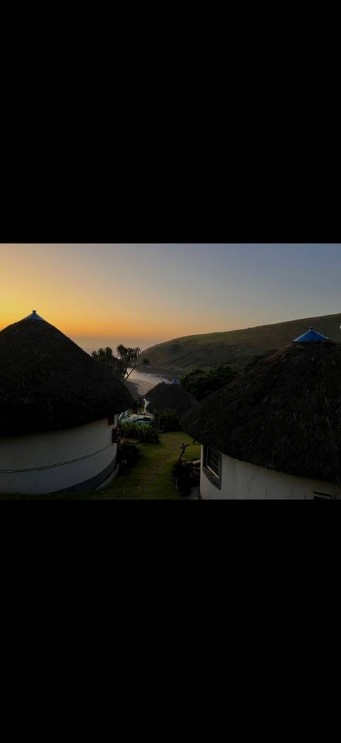 Traditional round thatched huts overlook a small bay at sunrise with mist rising over a green hillside.