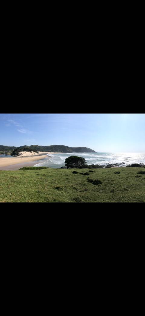 Wide coastal panorama of sandy beach, rolling waves and grassy headlands under a bright sky.