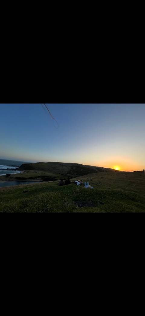 Group of friends sitting on a grassy hill watching the sun set over the coast and rolling waves.