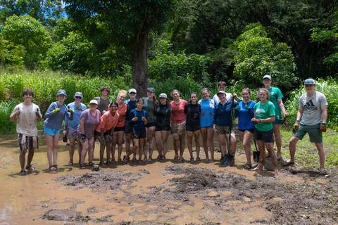 Large group of volunteers smiling after a muddy environmental project in a tropical clearing.
