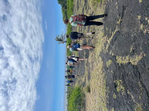 Line of hikers walking across rugged black lava fields toward the ocean under blue sky.