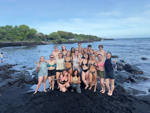 Large student group posing on black sand beach with blue ocean behind.