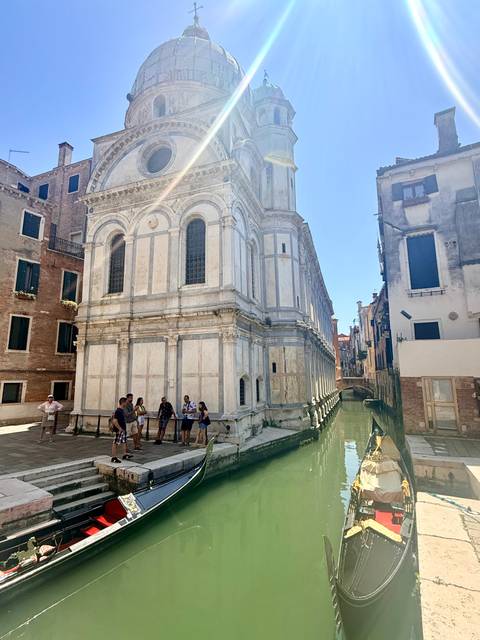 Travelers stand outside a marble-faced church beside a narrow Venetian canal.
