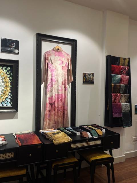 Interior gallery display featuring a pink silk dress, framed artwork and neatly arranged fabric items.