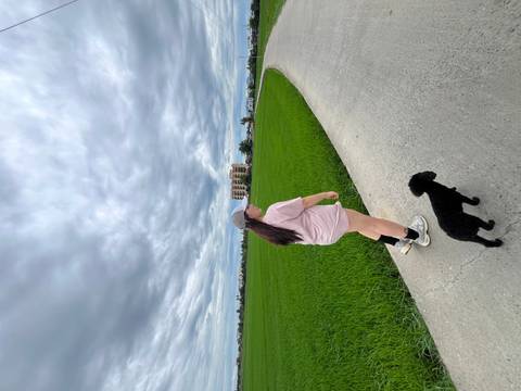 A young woman and her small black dog walk along a concrete lane beside vivid green rice fields under a cloudy sky.