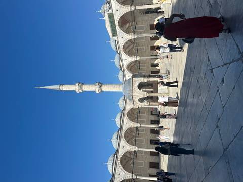 Visitors walk across the stone courtyard of an ornate Ottoman-era mosque dominated by a single tall minaret beneath a vivid blue sky.