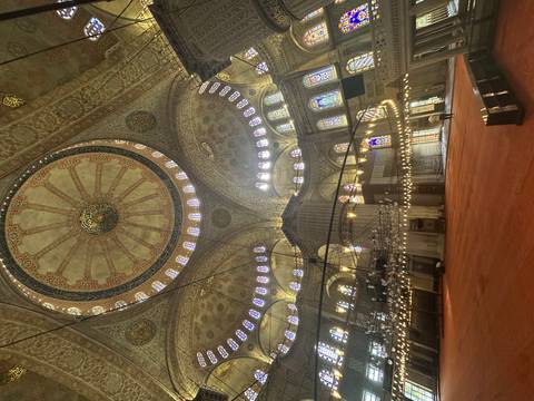 Lavishly decorated interior of a grand mosque with an enormous patterned dome, hanging chandeliers and stained-glass windows.