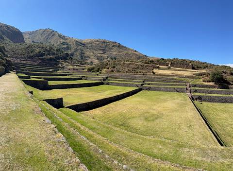 Terraced Inca ruins cascade down a green mountainside under a bright blue Andean sky.