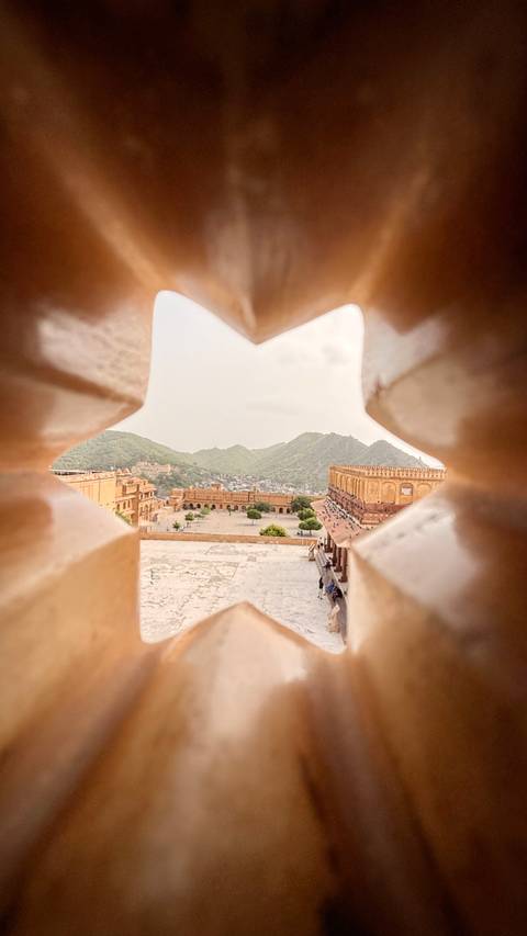 Creative peek through fortress lattice reveals Jaipur’s Amber Fort and surrounding green hills.
