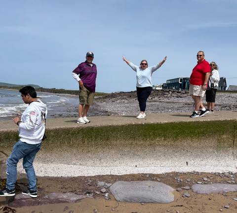 Smiling mature travellers pose playfully on a seaside concrete wall with shallow waves behind.