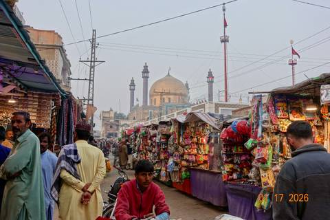 Busy Pakistani bazaar street with colorful stalls and mosque in background, date stamp visible.