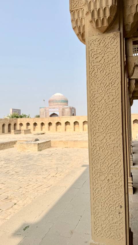 Detail of ornate sandstone carving on a pillar with a domed mausoleum and arched courtyard in the sun-baked background.