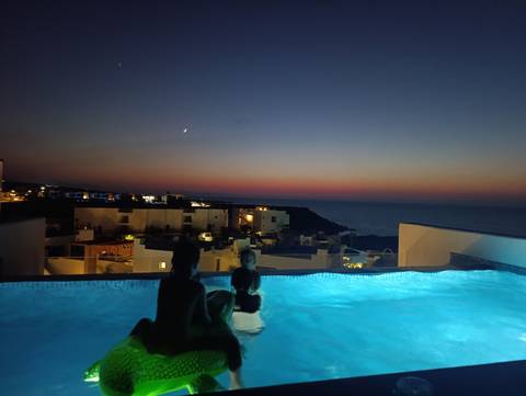 Silhouetted swimmers relax in illuminated infinity pool overlooking coastal town at dusk.