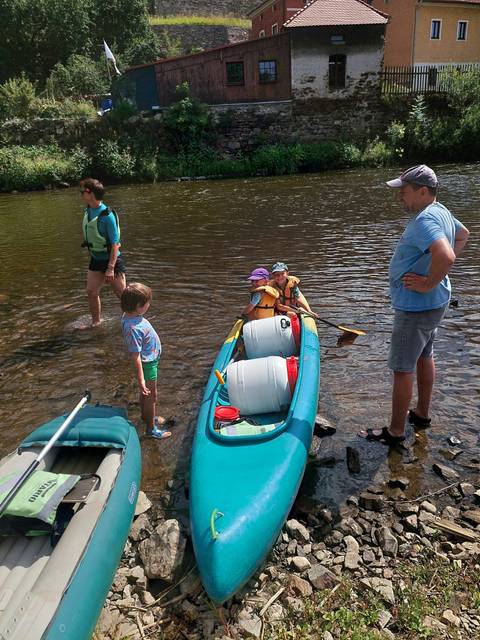 Family preparing canoe with barrels and gear in shallow river surrounded by greenery