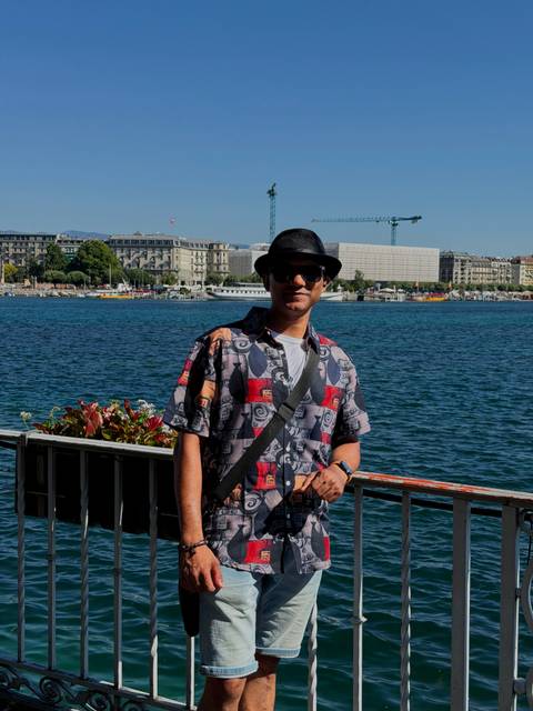 A man wearing sunglasses and a hat poses by the lakeside promenade with Geneva’s urban skyline and blue water behind him.