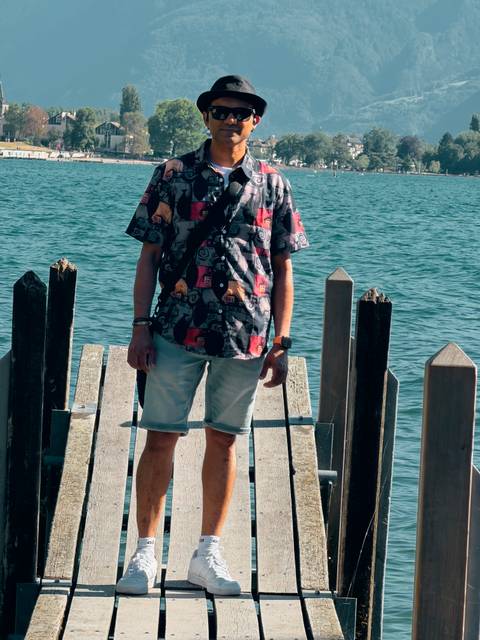 Lower-body portrait of a man standing on a wooden pier over a calm blue lake.
