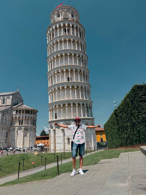 Traveller spreading arms wide in front of the Leaning Tower of Pisa under a clear blue sky.