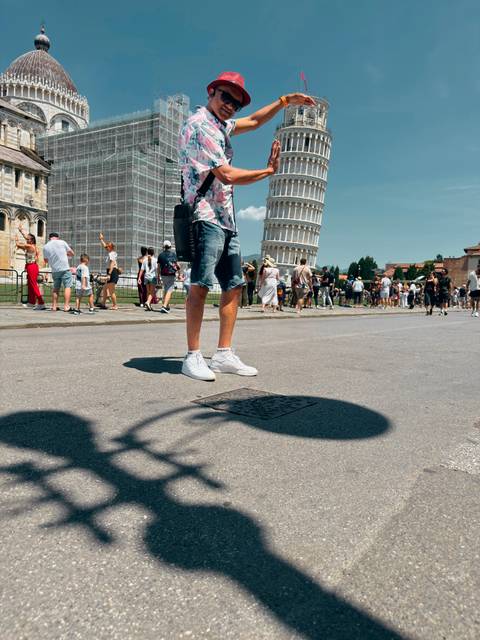 Ground-level photo featuring a traveller’s legs and shoes with the Leaning Tower of Pisa and crowds behind.