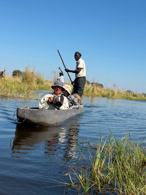 Traveller with telephoto camera rides a dugout canoe steered by a poler through the reeds of the Okavango Delta.
