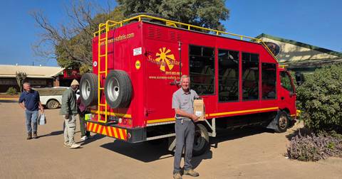 Bright red overland truck is loaded while travellers stand and smile in a sunny parking area.