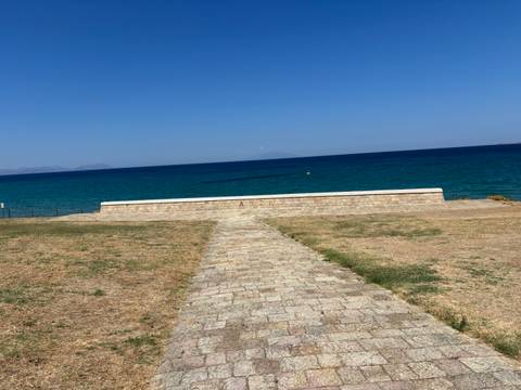 Stone path leading across dry grass toward a deep-blue sea horizon on a bright day.