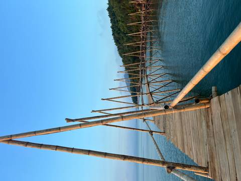 A rustic bamboo walkway supported by angled poles extends over turquoise sea toward distant green hills.