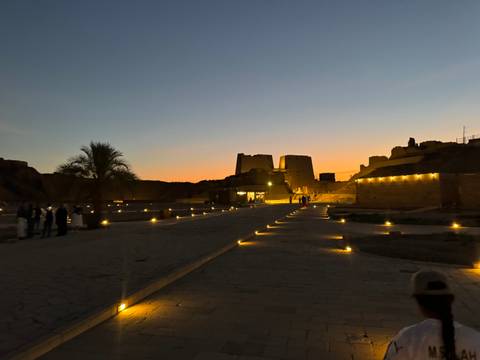 Temple complex subtly lit at dusk with a palm tree silhouette and visitors strolling the path.