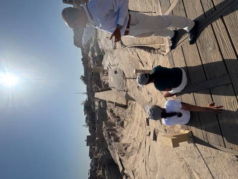 Visitors seated near the unfinished obelisk quarry in bright sunlight while guide explains.