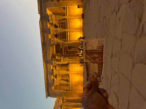 Blurry foreground hand holding a 50-pound note against a lit temple facade at dusk.