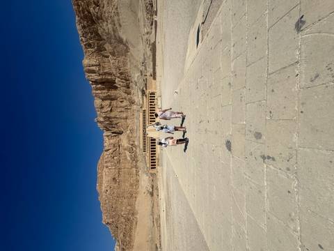 Tourists walking toward Hatshepsut Temple with towering cliffs under a deep blue sky.