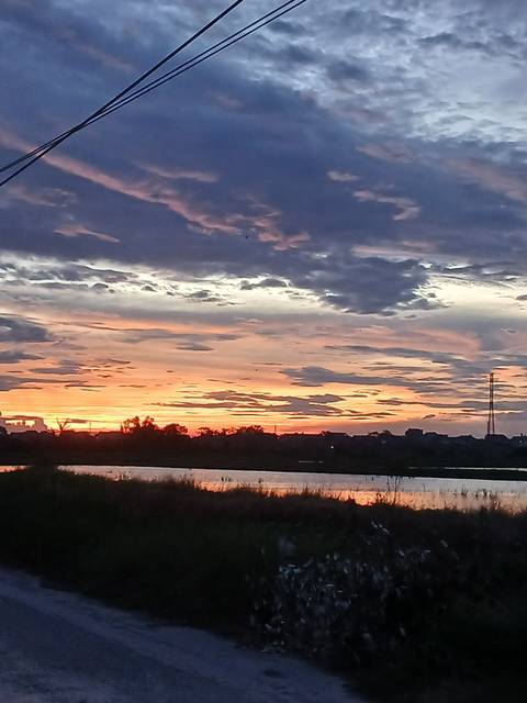 Colourful sunset with dramatic layered clouds reflected on a calm river beyond a distant village silhouette.