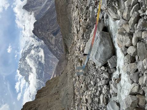 Rainbow-planked suspension bridge crossing a rocky glacial stream with soaring snowy peaks beyond